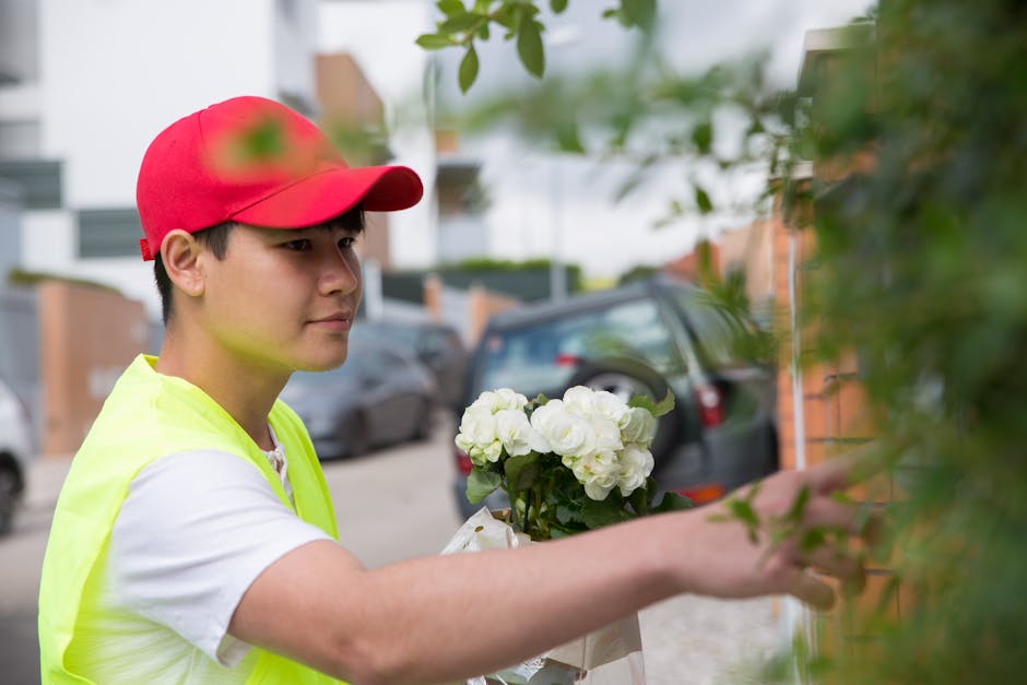 A young male flower delivery person wearing a red cap and a neon yellow vest is seen outdoors, holding a bouquet of white hydrangeas wrapped in white paper. He is reaching out with his right hand to p